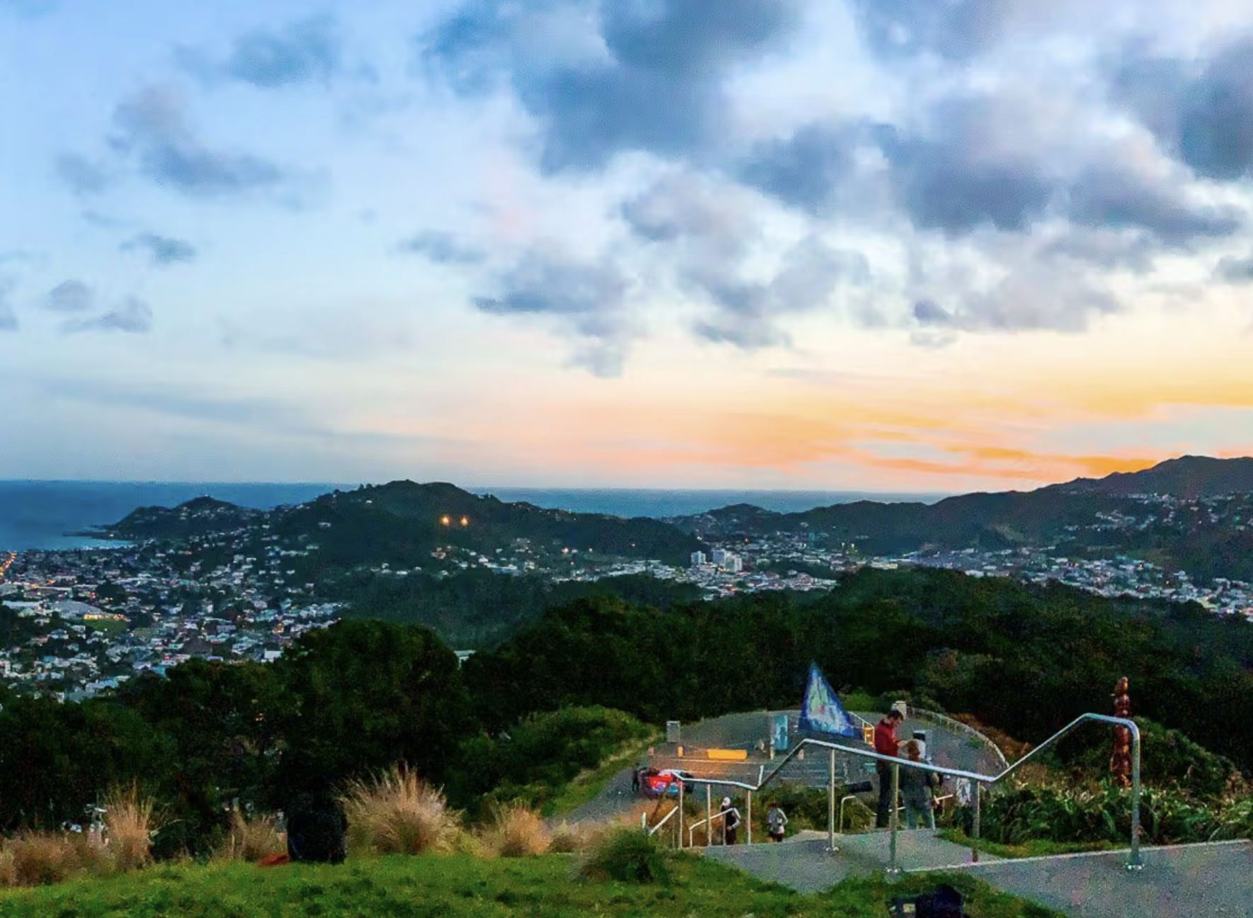 Viewpoint overlooking a coastal city at sunset, with hills, sea, and a few people standing by the railing on a stairs path.